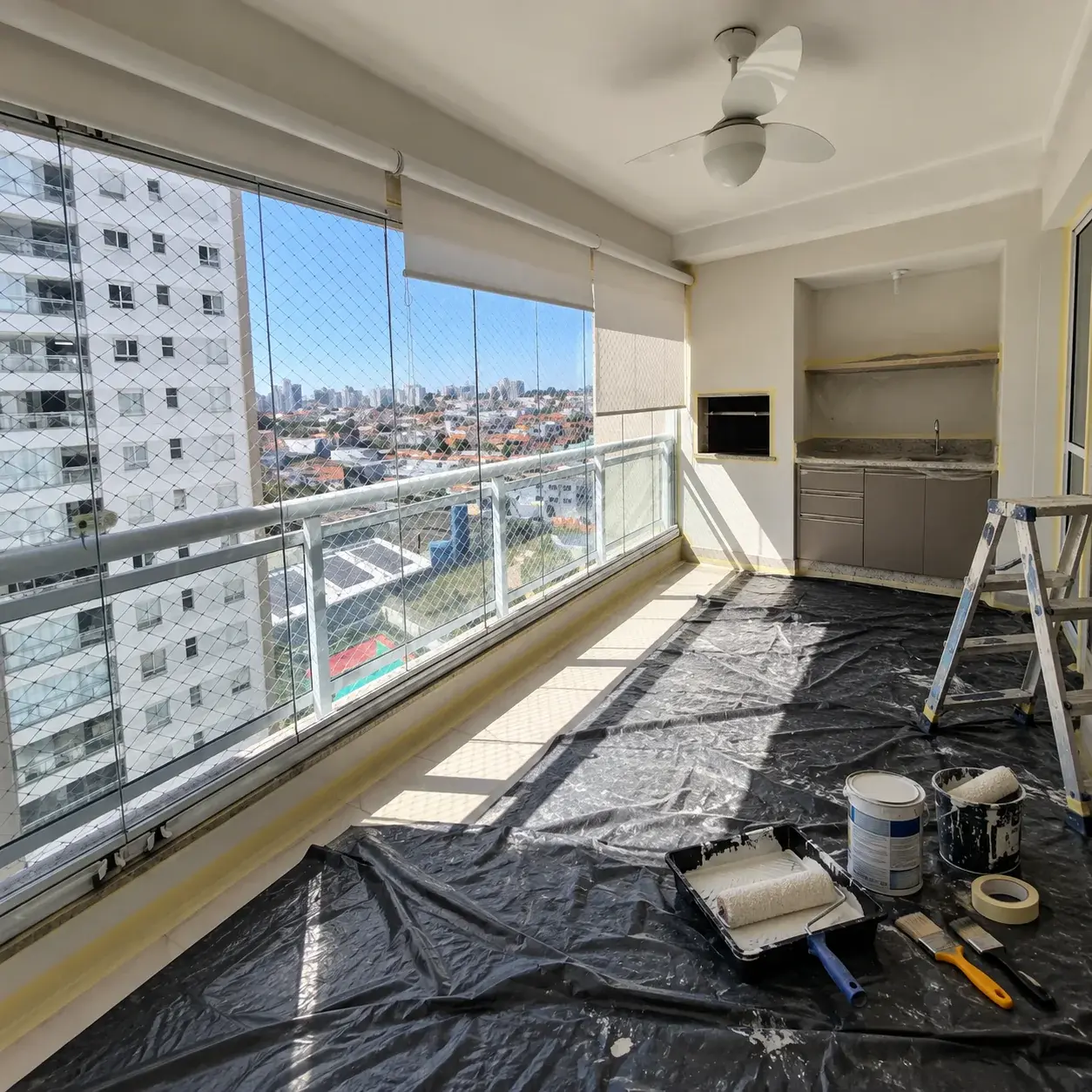 Varanda de apartamento em pintura, com o piso protegido por lona, rolos e latas de tinta